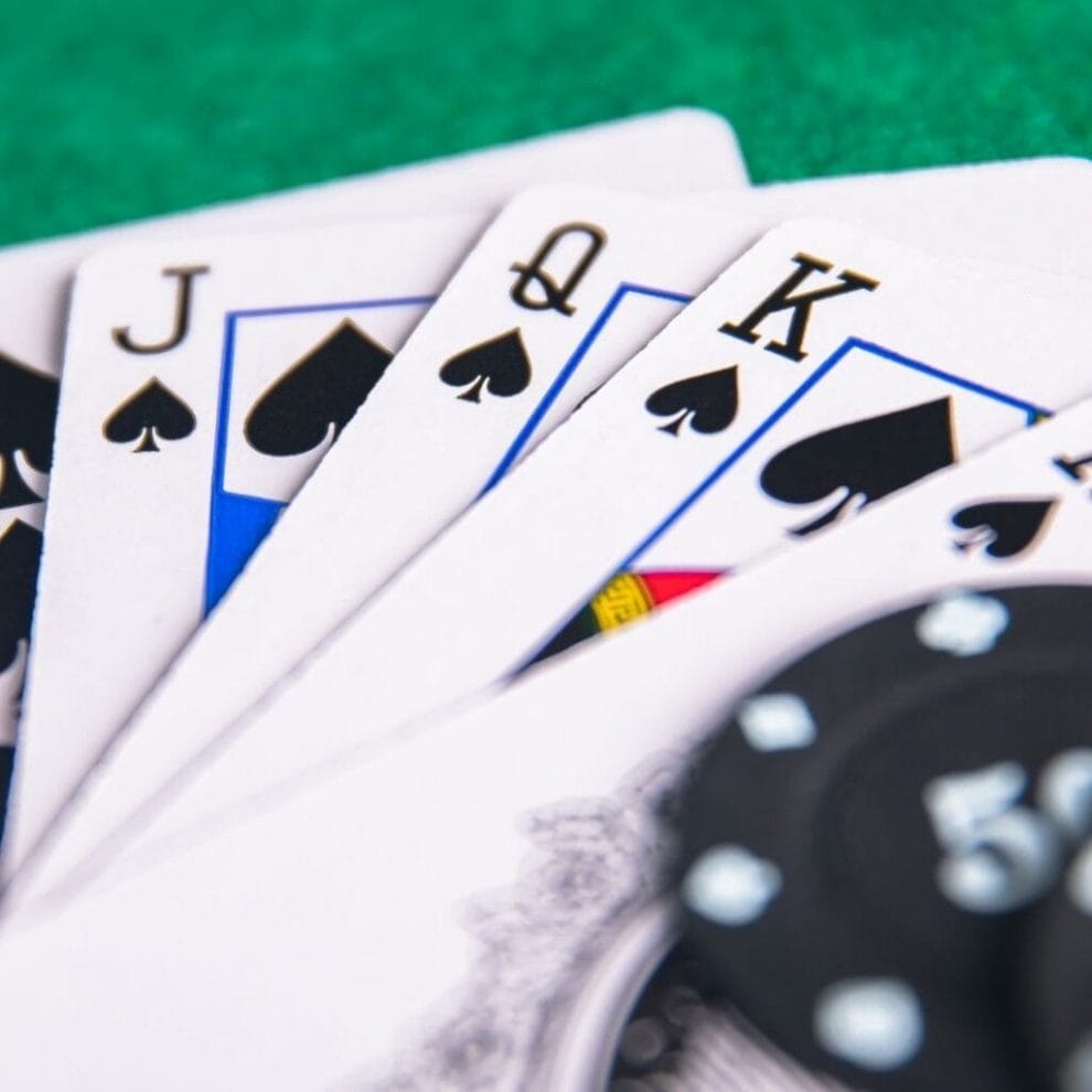 close up of a royal flush of spades fanned out on a green felt poker table with a blurred black poker chip on the cards in the foreground