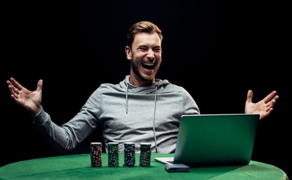 A poker player laughs with their hands spread out. A laptop sits on a green table next to four stacks of poker chips and a deck of cards.