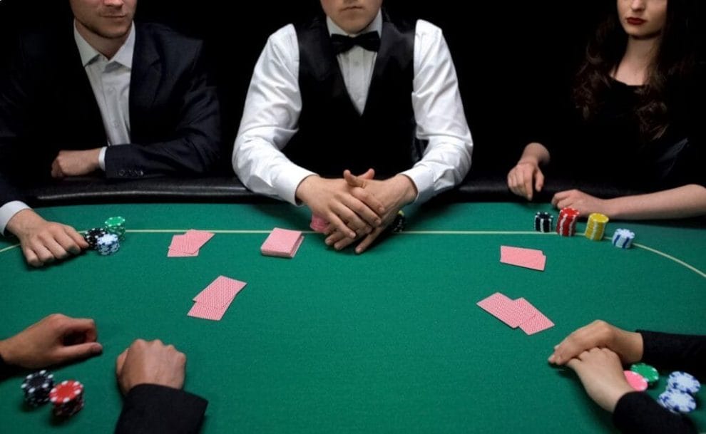 Four poker players seated around a table with a croupier in the middle. Stacks of poker chips and scattered playing cards.