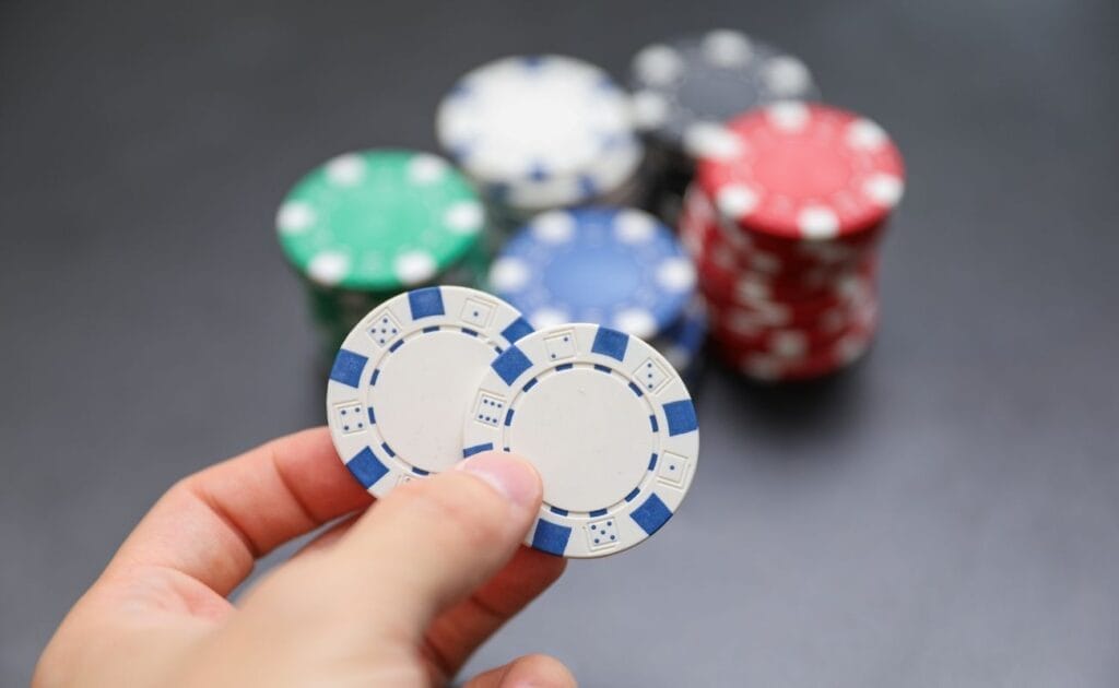 Hand holding two poker chips, with five stacks of poker chips in the blurred background.