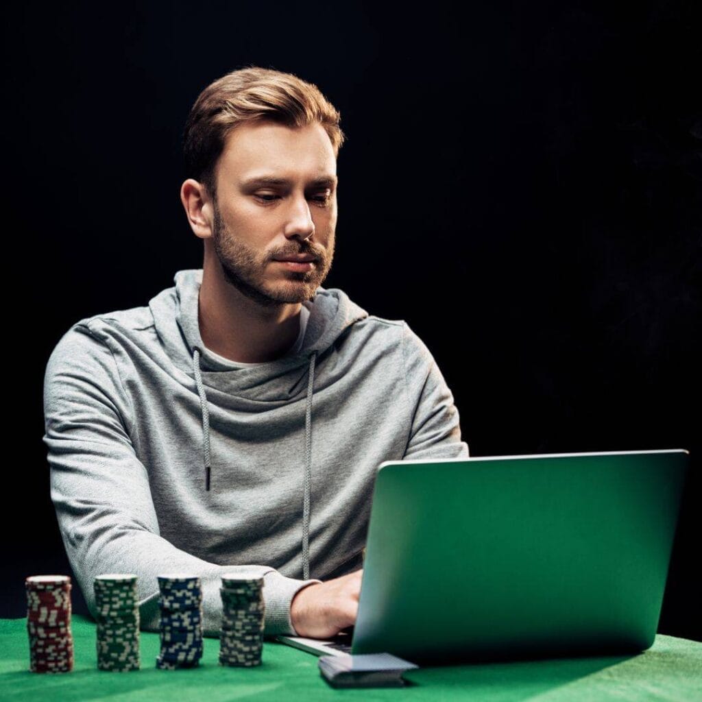 Man gambling online on his laptop, set on a green felt cloth. Four stacks of poker chips and a blue deck of cards beside him.