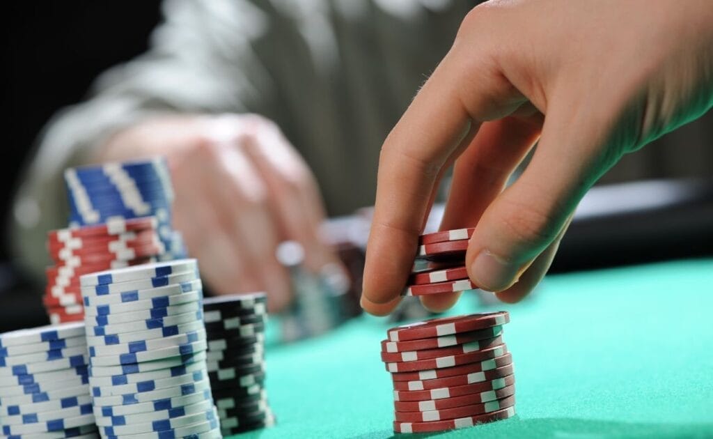 Hand lifting four poker chips from a stack, with a player on the opposite side of the table and additional chip stacks to the left.