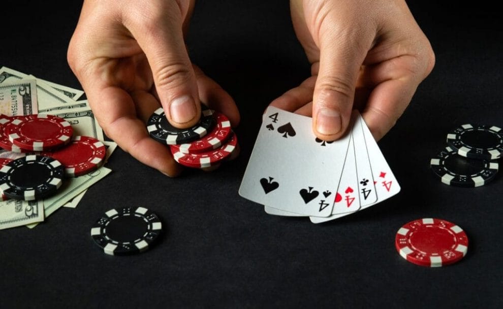 A poker player holds a few chips in their left hand and 4 fours in their right hand. There are poker chips and cash on the table beneath their hands.