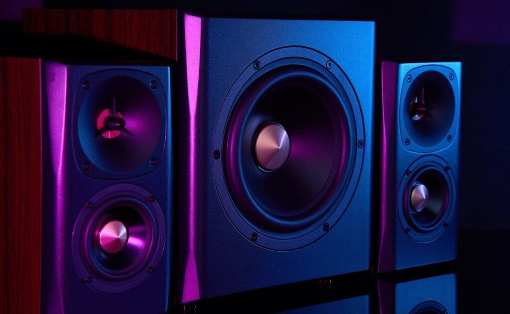A pair of speakers on either side of a subwoofer sitting on a reflective black table. The speakers and subwoofer are bathed in neon light.