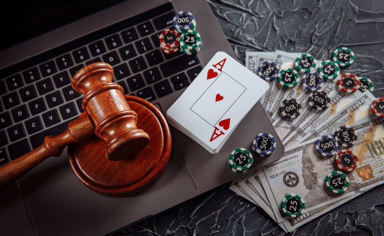A judge’s gavel and sound block on top of a laptop with playing cards, cash, and casino chips surrounding them.