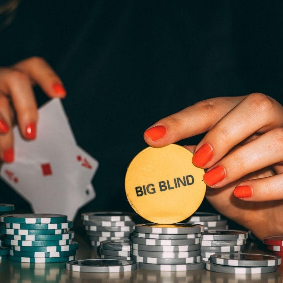 A person holding a “Big Blind” chip, on top of silver and green poker chips, with two aces in their other hand.