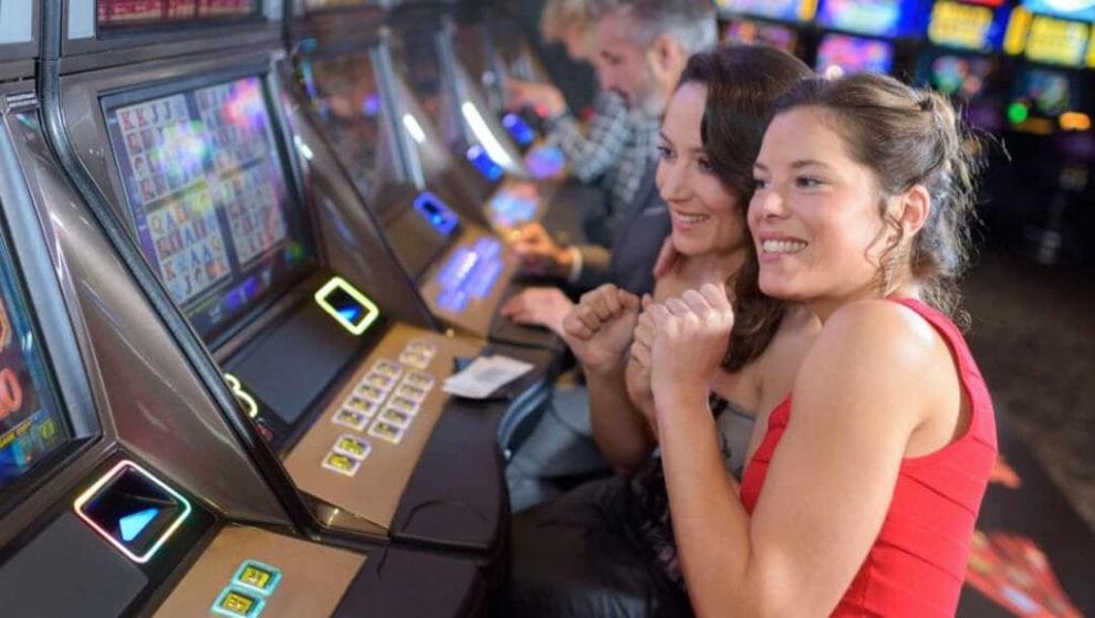 Two friends lean together while watching reels spin on a slot machine in the casino.