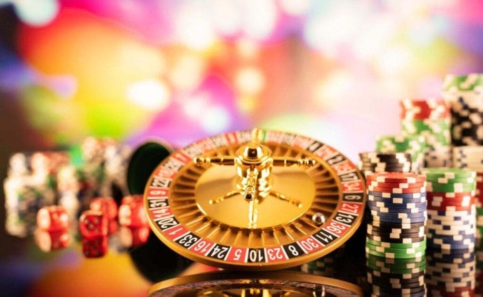 A small gold roulette wheel on a reflective table with casino chips and dice surrounding it. 