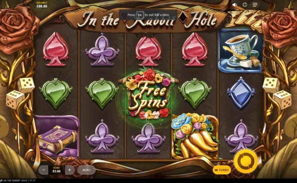 In the Rabbit Hole reels with dice and roses set as the reel frame against a brown-hued background. The reels are filled with playing card suits such as blue diamonds, green hearts, red spades and purple clubs alongside the Free Spins symbol, a teacup and a book.