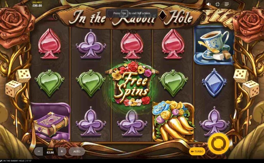 In the Rabbit Hole reels with dice and roses set as the reel frame against a brown-hued background. The reels are filled with playing card suits such as blue diamonds, green hearts, red spades and purple clubs alongside the Free Spins symbol, a teacup and a book.