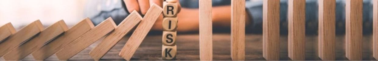 A risk management concept image of a man using his finger to stop wooden blocks that spell out “risk” from falling like dominos.