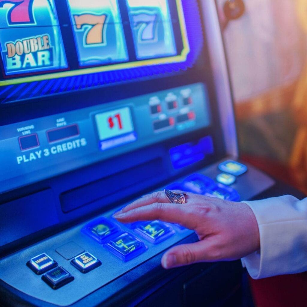 A person sitting at a slot machine inside a casino, with their hand about to push a button on the slot machine.