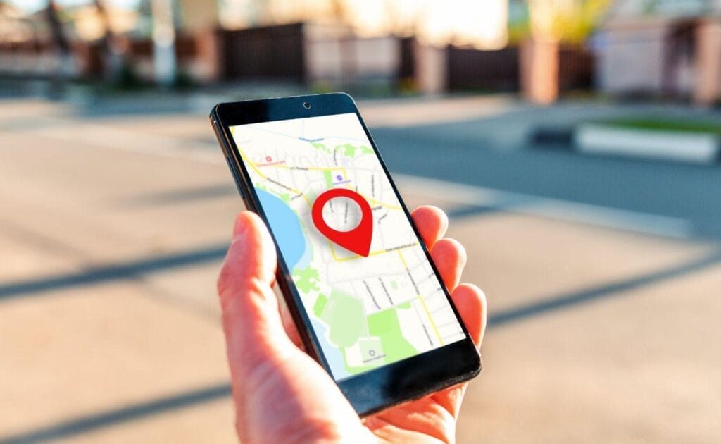 A close-up of a hand holding a cellphone with a map and red location pin on the screen; A blurred street is in the background.