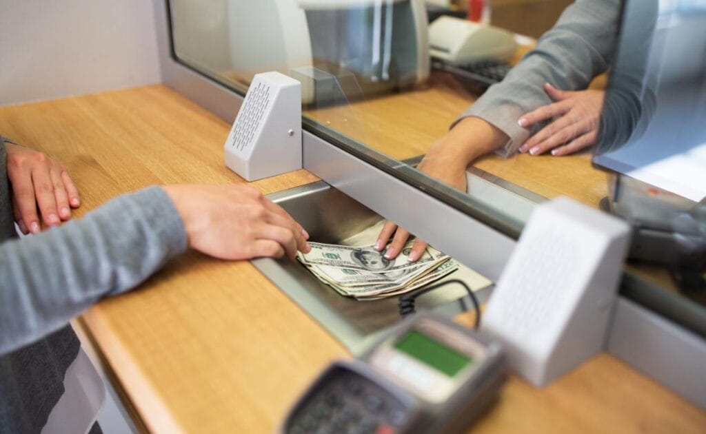A person passing money through a bank teller with hands receiving it on the other side of the window.