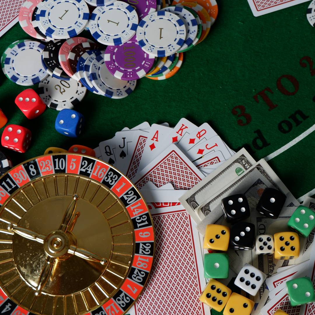 A range of casino game equipment sitting on a casino game table, including a mini roulette wheel, dice, playing cards, and casino chips.