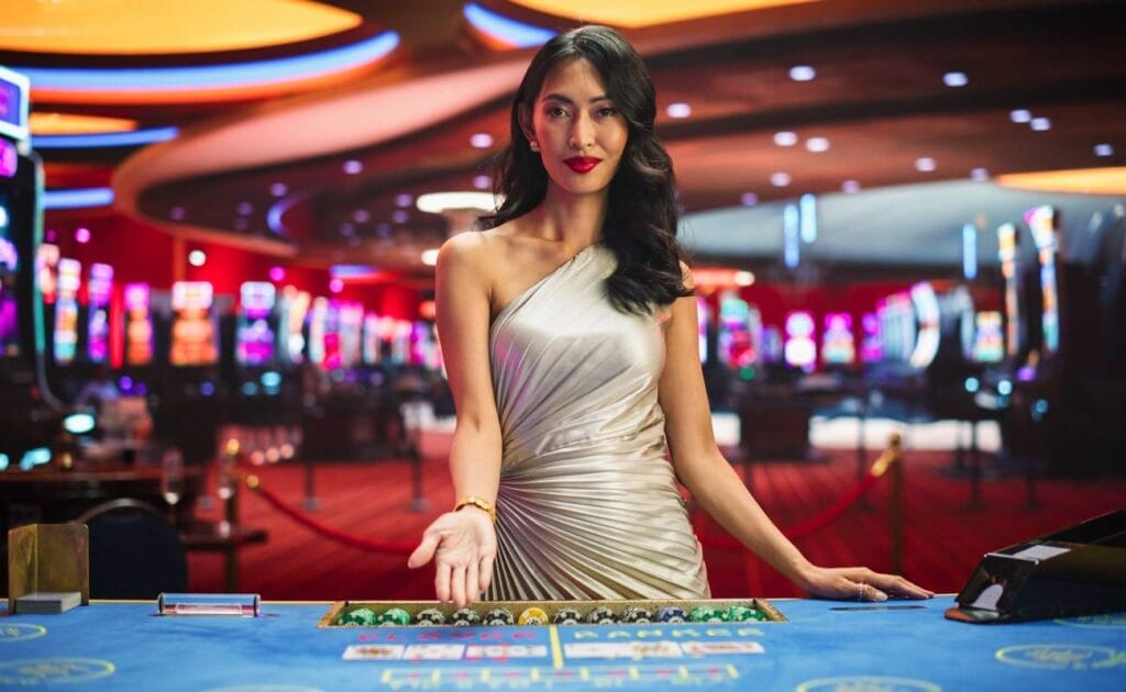 A woman dealer in a silver dress gesturing to play at a baccarat table in a casino.