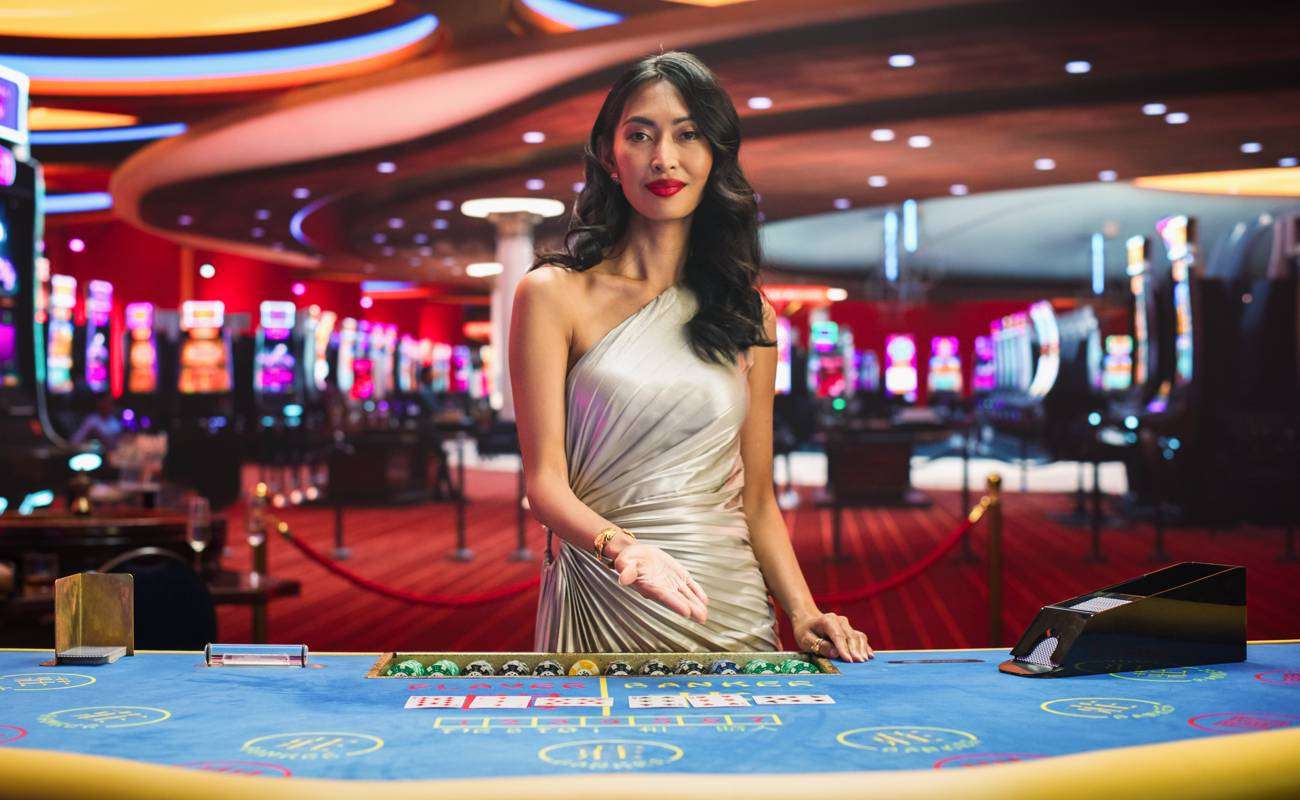 A female casino dealer in a silver dress gestures toward a baccarat table.