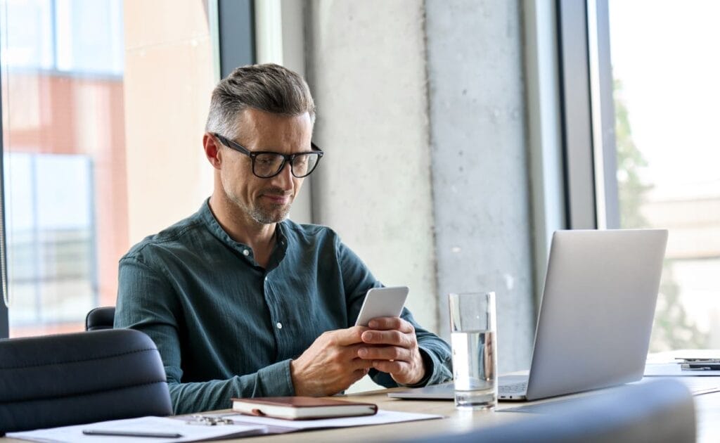 A man sitting at a desk with a laptop, a journal, and a glass of water. He is wearing glasses and holding a smartphone.