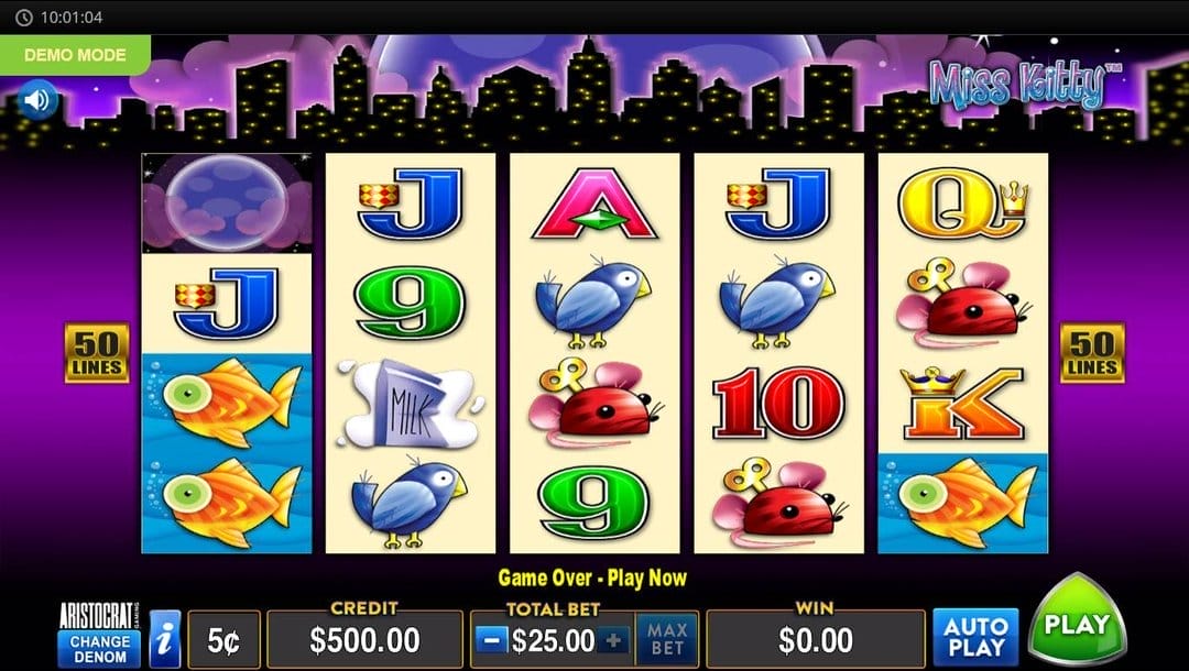 A screenshot of the slot reels in the Miss Kitty online slot game. The graphics are in a bright, cartoonish style. The background features a stylized purple city skyline glowing with yellow-lit windows beneath a night sky scattered with stars. The top-right corner displays the game title Miss Kitty in a playful, bubble-like font. In the foreground, five vertical reels are filled with colorful, animated symbols like goldfish, milk cartons, bluebirds, wind-up mice, and the standard card icons J, Q, K, 10, and 9. The reels are set against a pale cream background framed by a rich purple border, while yellow “50 LINES” labels appear on both sides of the grid. The game interface is below the slot reels.