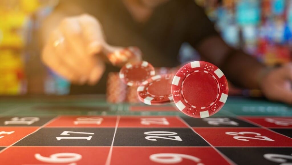 Chip kasino merah terbang di udara menuju meja roulette selama pertandingan, dengan pemain yang bergerak dan latar belakang kasino yang kabur.