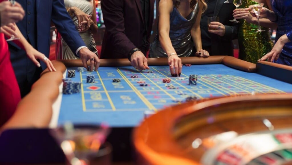 Pemain kasino berpakaian bagus menempatkan taruhan di atas meja roulette biru di lingkungan game yang glamor, dikelilingi oleh minuman dan energi kehidupan malam yang semarak.