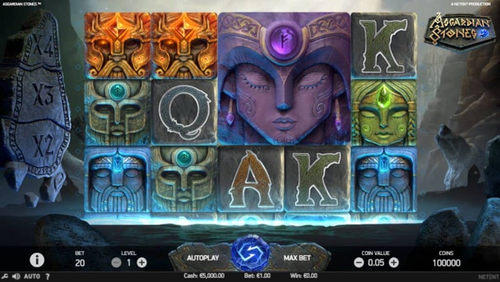 A screenshot of the Asgardian Stones slot reels. The background shows a river flowing between two mountain edges. In the foreground, the slot reels with various Norse-themed stone faces, as well as playing card symbols. A sculpture with multipliers is visible on the left of the reels.