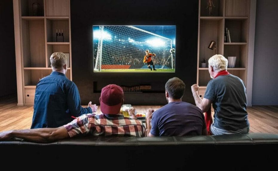 Rear view of a group of friends sitting on the couch watching soccer on a large TV screen at home
