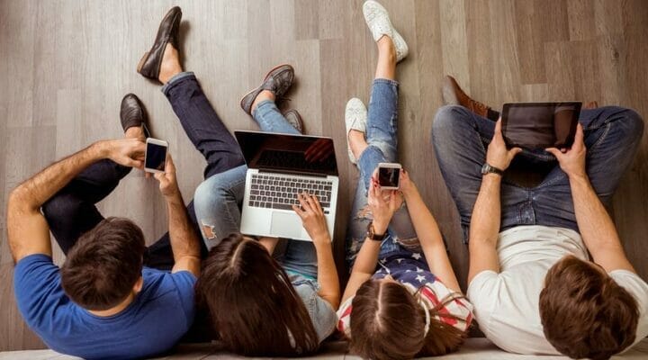 A group of people sitting on the floor using a laptop, tablet PC, and smartphones.