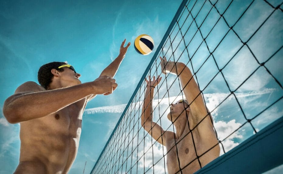 A male beach volleyball player attempts to hit the ball over the net while an opponent tries to block the ball.