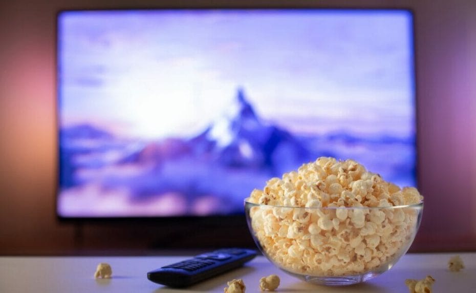 A bowl of popcorn on a coffee table with a movie on the TV screen in the background.