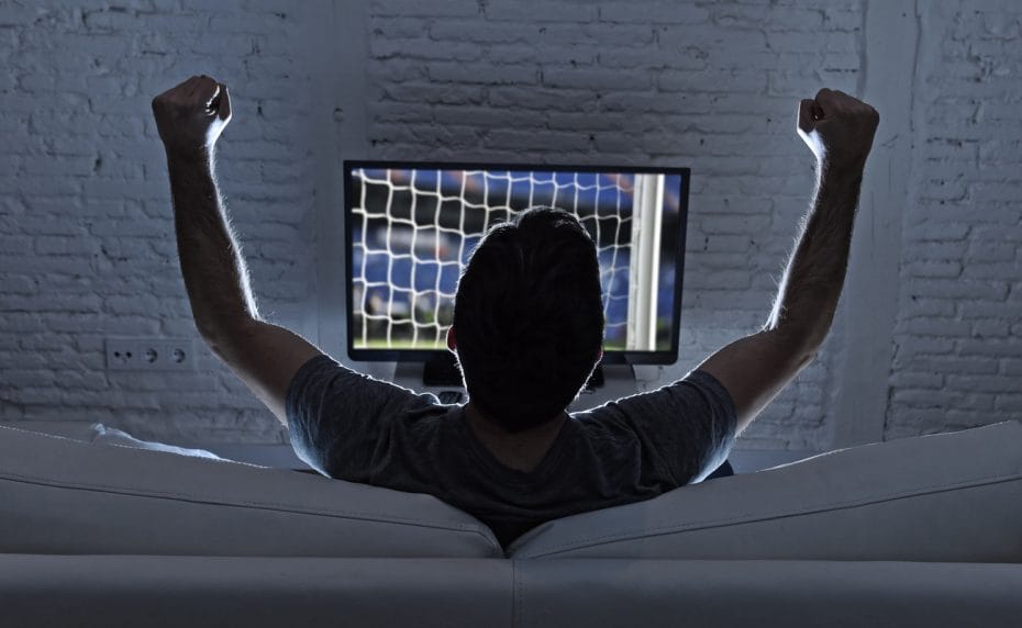  The back view of a man watching soccer on the couch with his arms in the air.