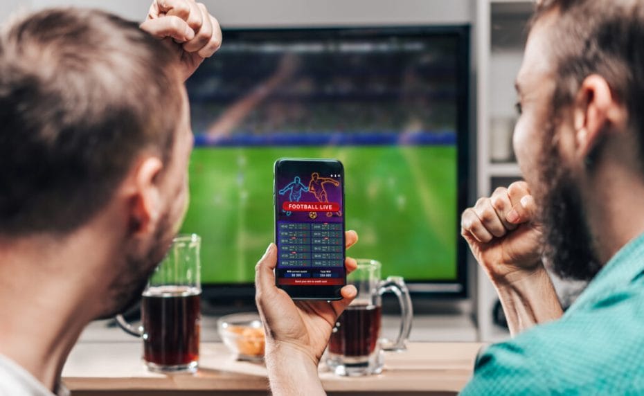 Two male friends watching soccer on the TV and in-game betting on a mobile phone.