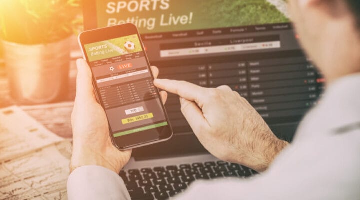 A person holds a phone in front of a laptop with live sports betting on the screen.
