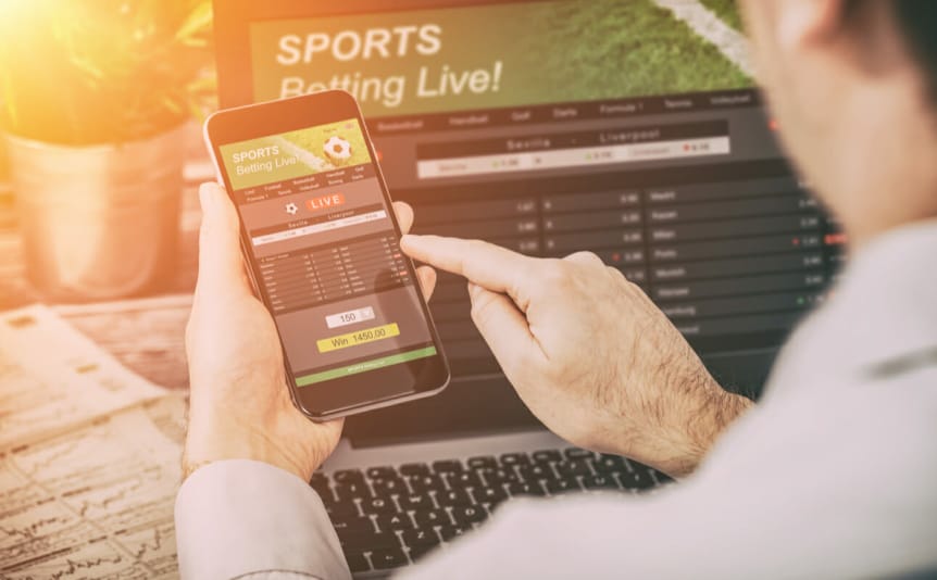 A person holds a phone in front of a laptop with live sports betting on the screen.