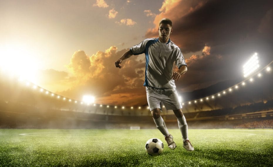 A soccer player with the ball and stadium lights behind him.