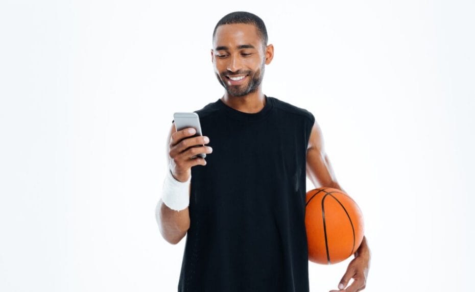 A young man carrying a basketball smiles while he looks at his smartphone.