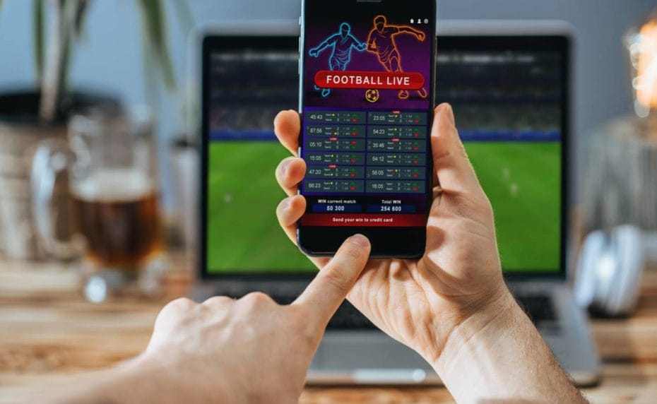 A man places a soccer bet on his phone while watching soccer on his laptop.