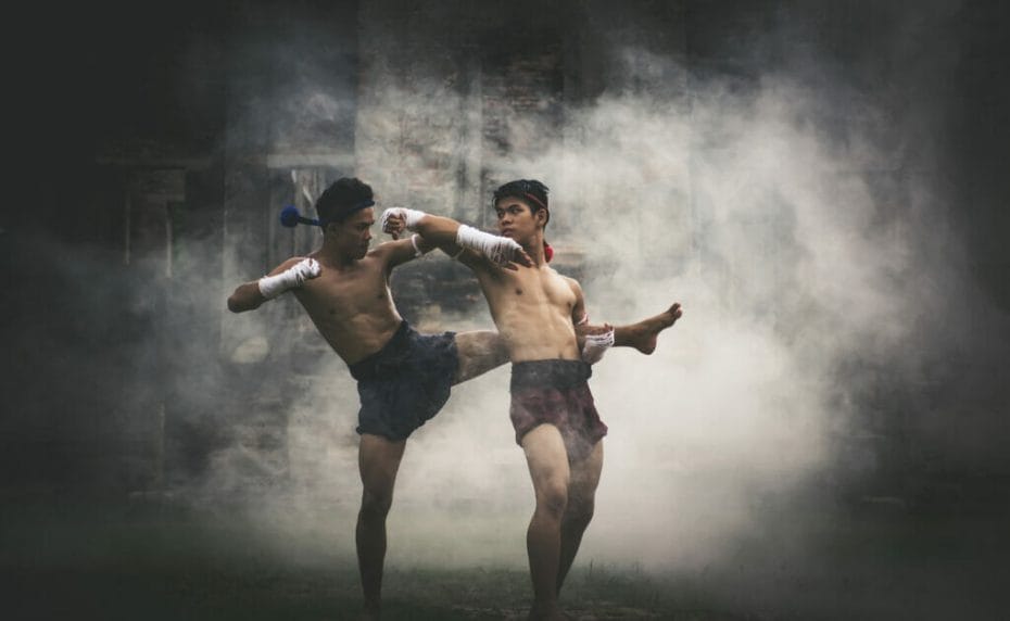 Two men practice Muay Thai outdoors with mist behind them.