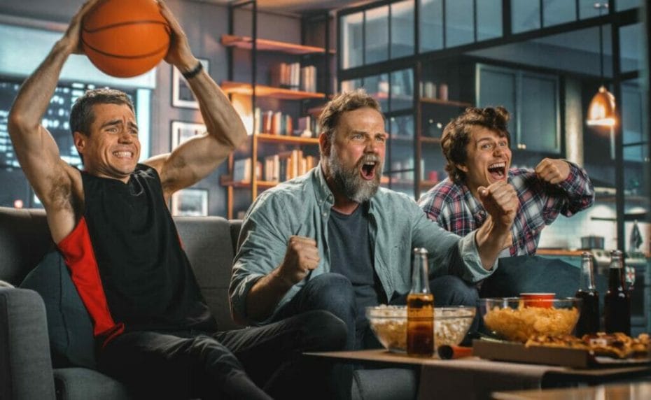 Three men sitting on a coach enthralled by what they are watching. One of them holds a basketball in their hands.