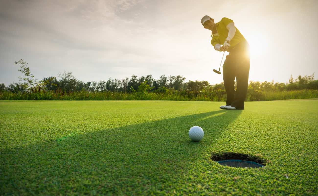 A golfer putts the ball towards the hole with the sun behind him.