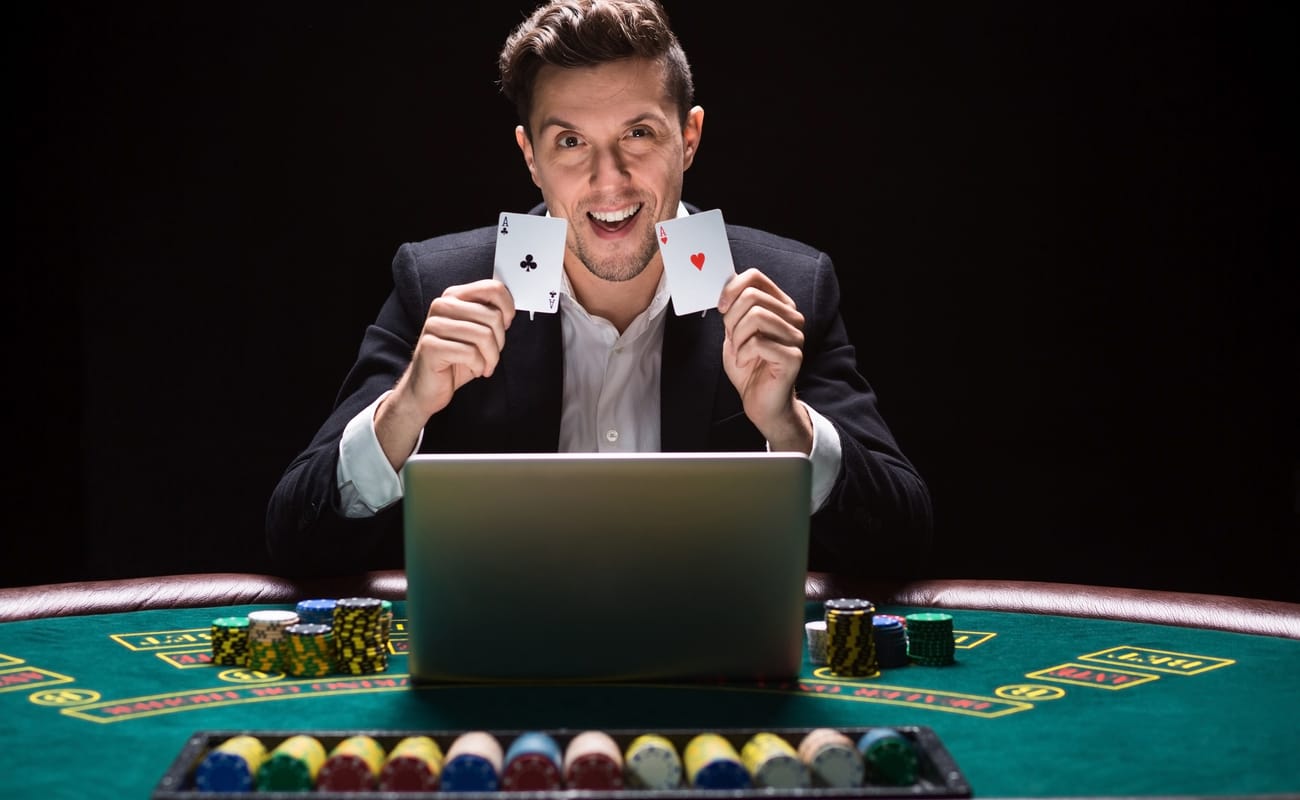 Online poker player holding playing cards and sitting at a casino table with his laptop and casino chips. 