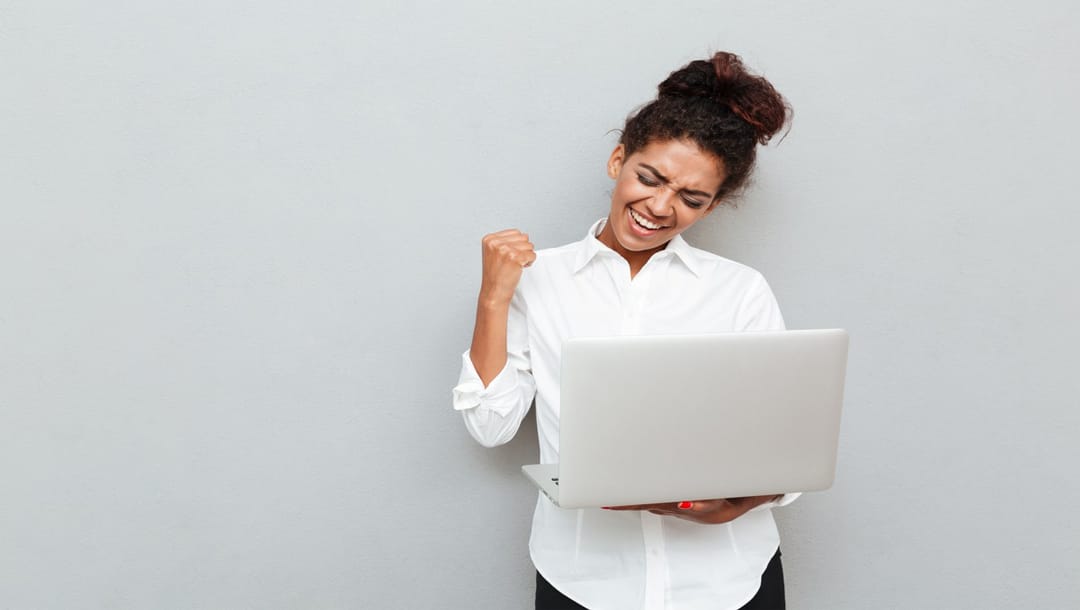 Photo of a cheerful businesswoman standing against a grey wall, holding a laptop, and celebrating a win.