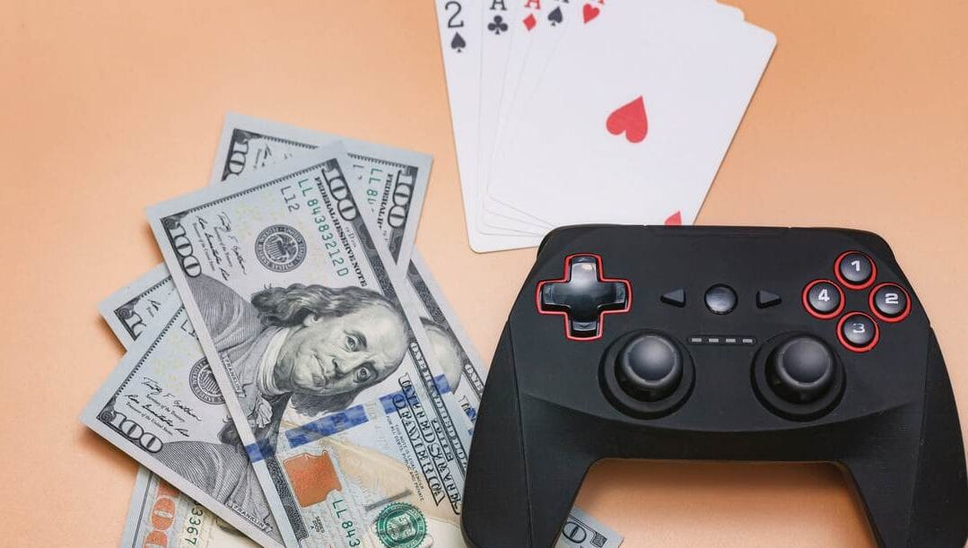 Game console, 100 dollar bills and playing cards against a peach background.