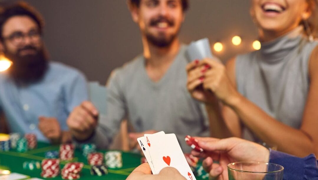 A man’s hands holding two poker cards and a chip with people smiling in the background.