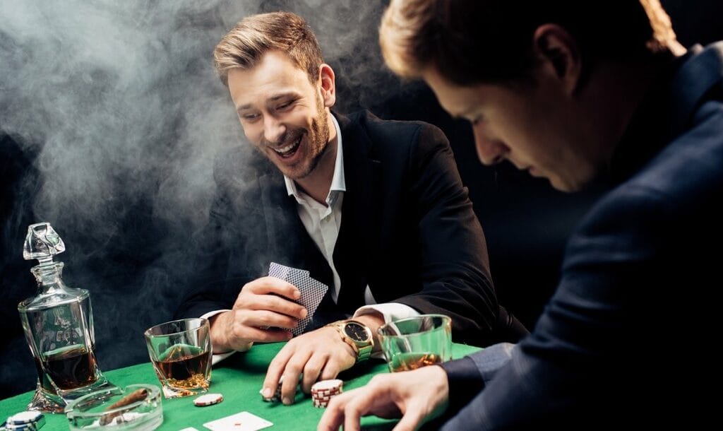 Two poker players drink whiskey and laugh while at a poker table.