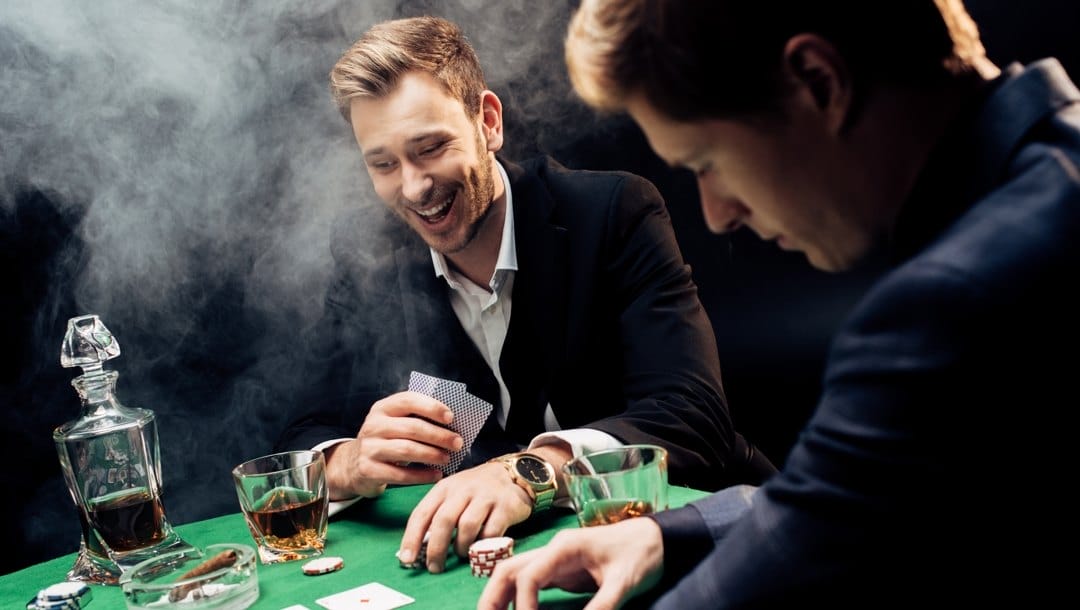Two poker players drink whiskey and laugh while at a poker table.