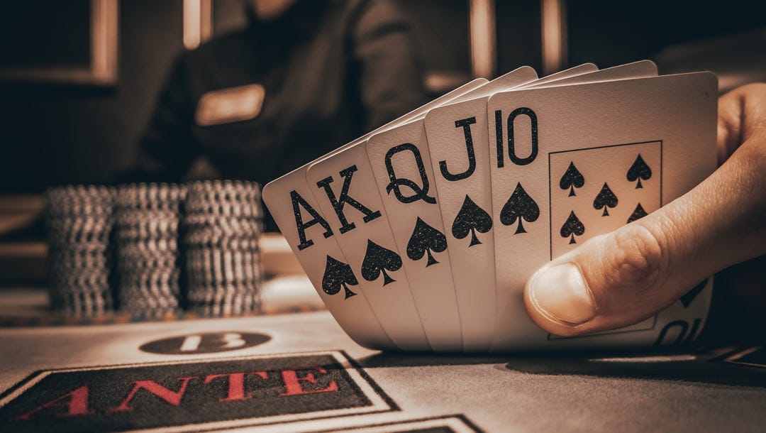 A hand holding a royal flush at a poker table with casino chips in the background.