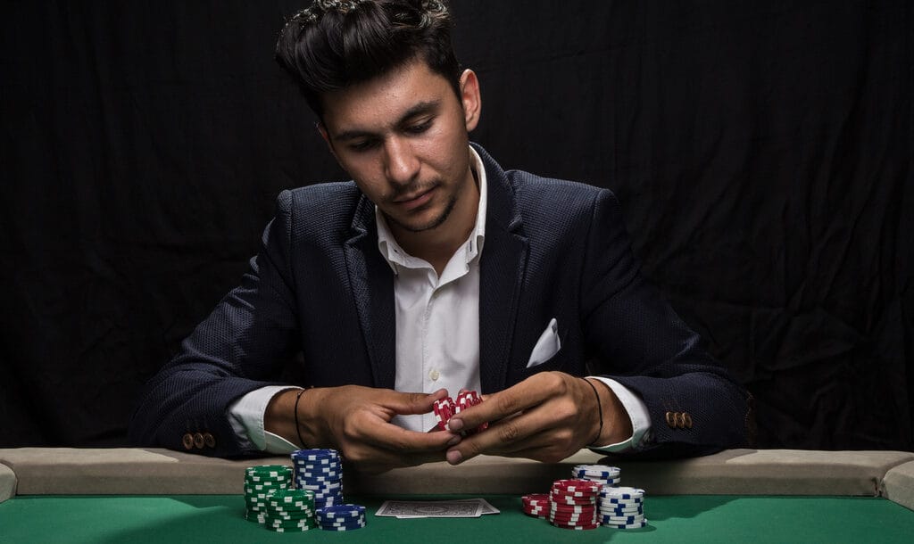 A serious-looking man in a suit sits at a poker table with chips in his hand.
