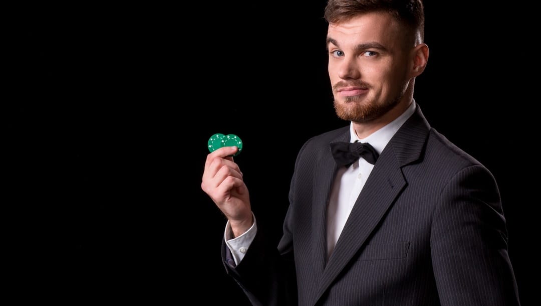A man in a suit and bow tie holds two green poker chips between his fingers.