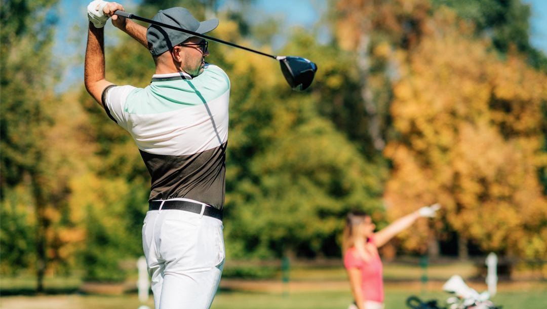 A golfer strikes the ball with another player in the distance.